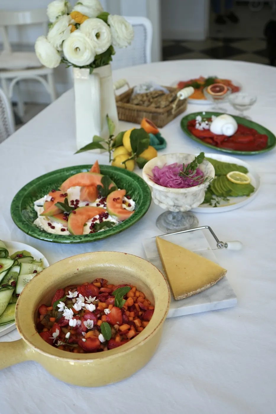 A white table with melon, burrata, fruit salad and fresh flowers